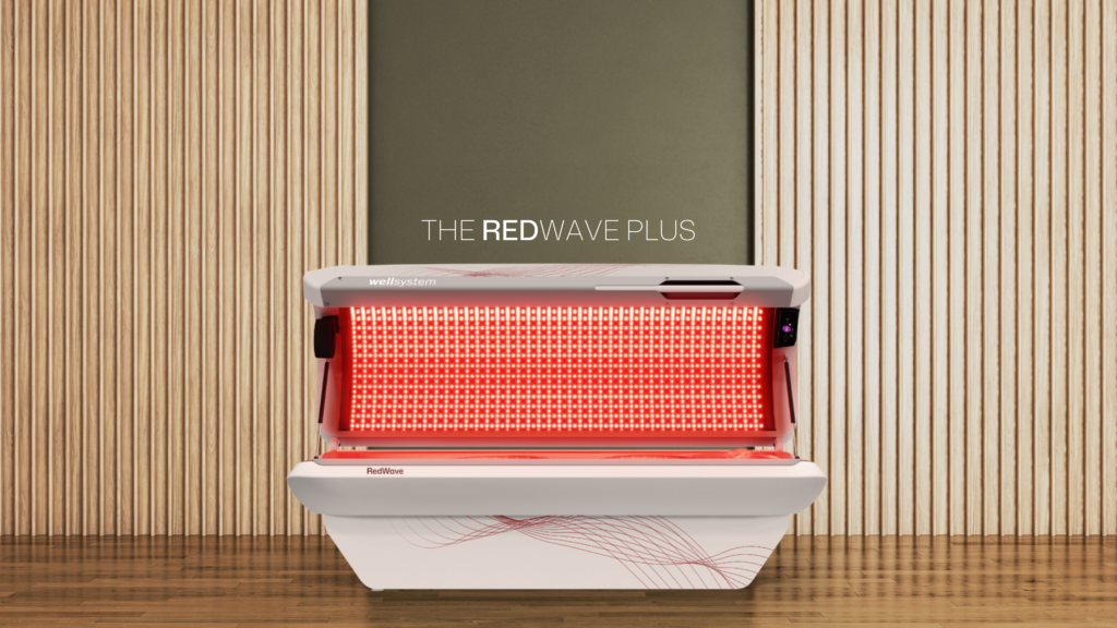An image of JK Products and services new addition to the Wellsystem brand; the RedWave Plus unit. A full Red Light Laydown device for wellness, sits in a calming environment consisting of warm wood walls and an olive green backdrop that lets the red glow of the unit pop.